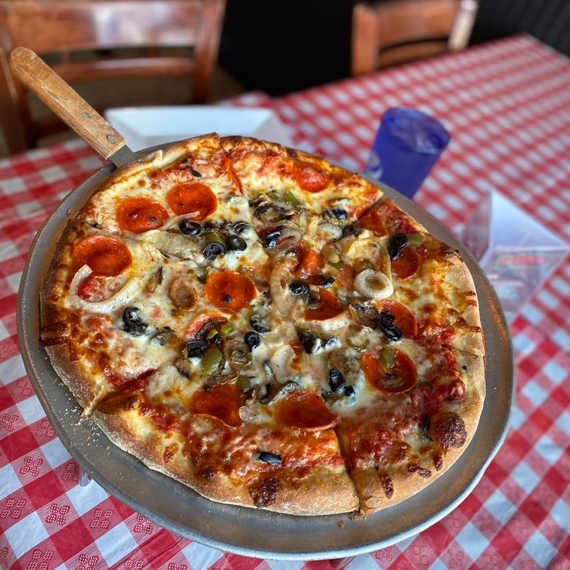 Pizzitalia's pepperoni, olive, mushroom, and onion pizza on a metal pan with a red and white checkered tablecloth.