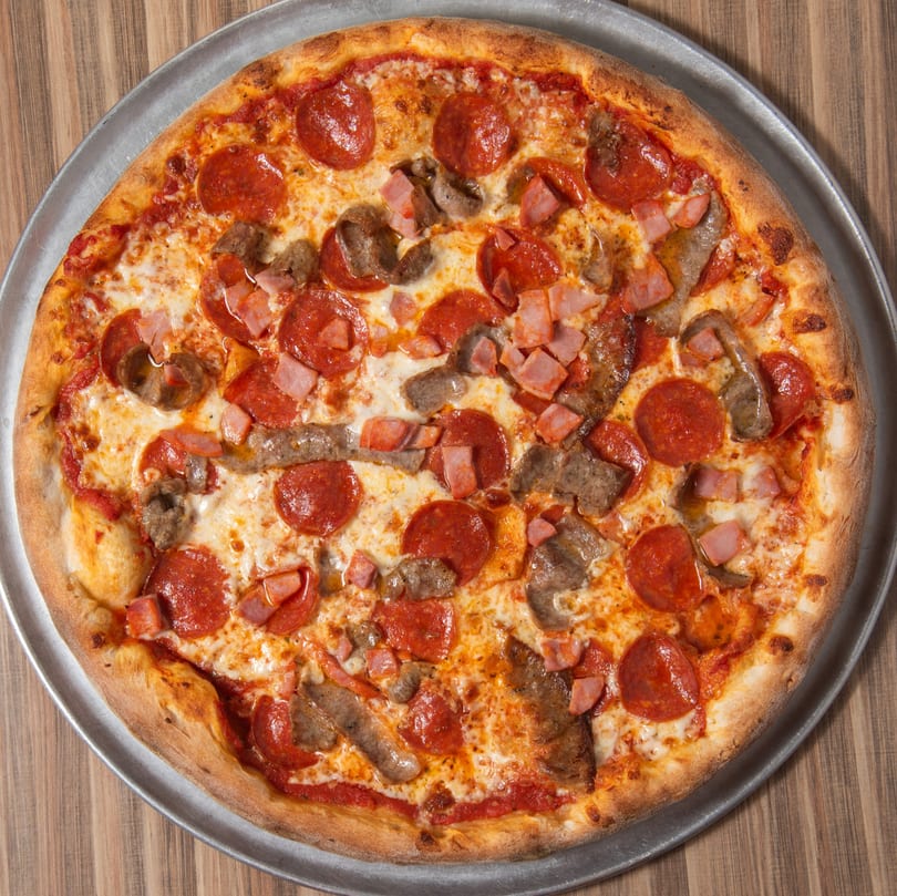 A whole meat lover's pizza with pepperoni, ham, and sausage on a metal pan.