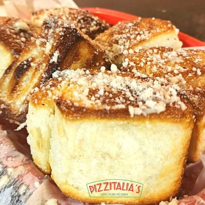 Close-up of golden-brown garlic knots from Pizzitalia's, sprinkled with Parmesan cheese in a red basket.